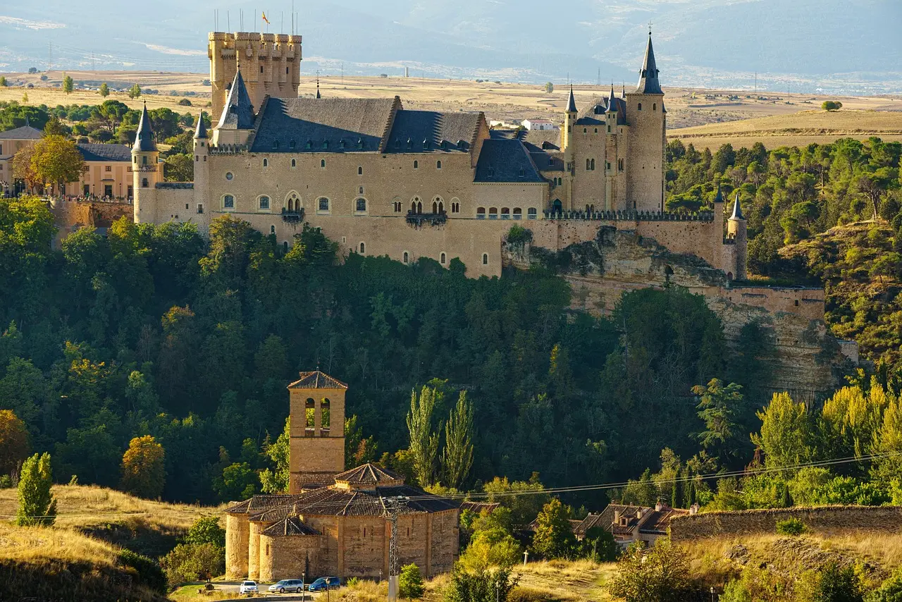 The Alcázar of Segovia at dawn — its fairytale silhouette mirrored in tranquil waters, capturing the romance and regal legacy of must-visit places in Spain. Alcázar of Segovia at sunrise, reflected in a calm river with mist rising