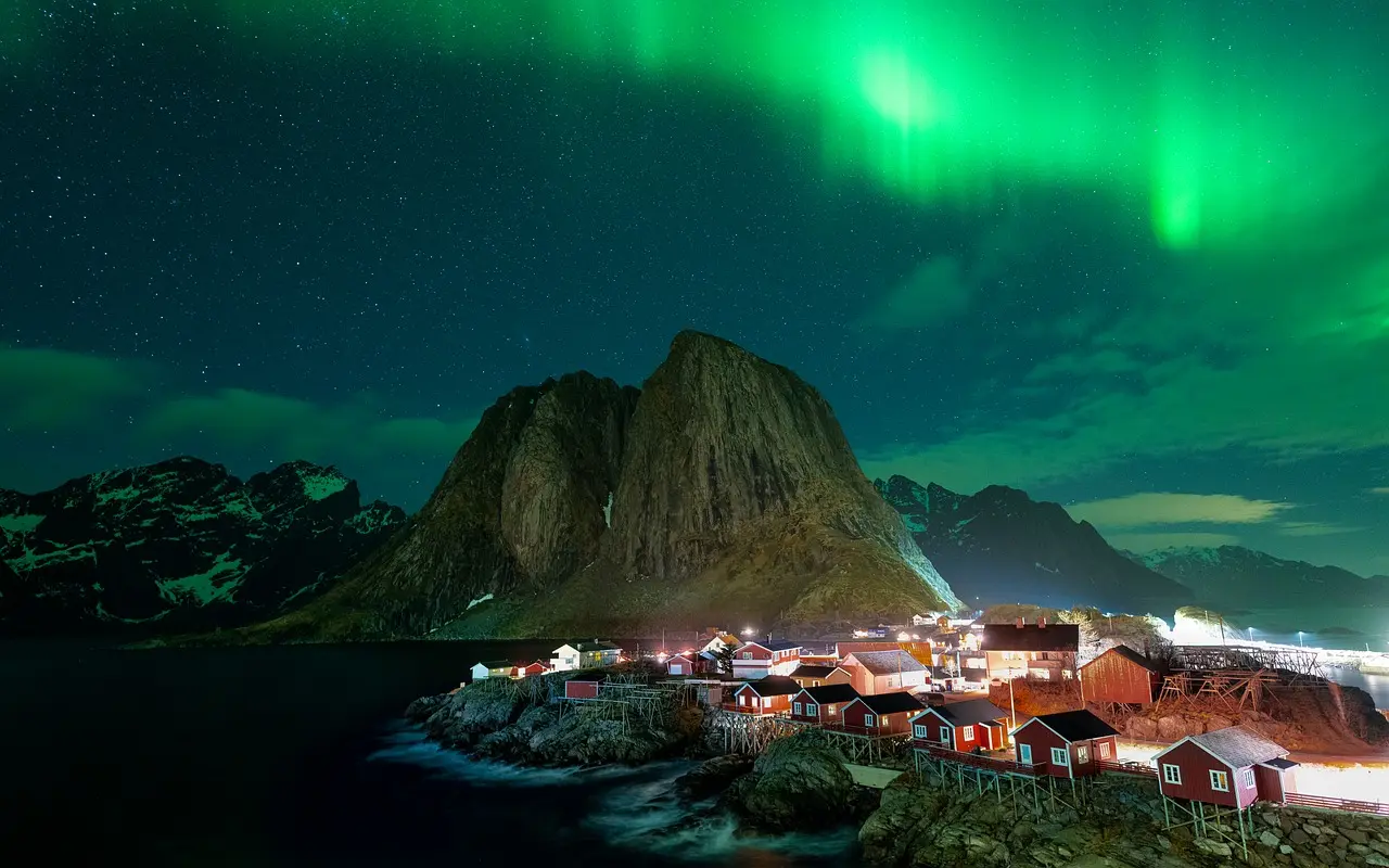 A historic fisherman’s cabin in Lofoten under the northern lights—embodying the minimalist, nature-immersive ethos of relaxation travel. Traditional red rorbu cabin on wooden stilts beside a still fjord under green aurora lights
